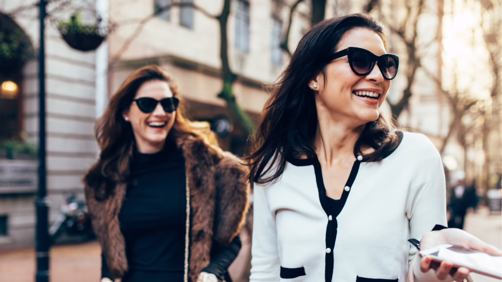 Two women walking on a city street, both wearing sunglasses and smiling.