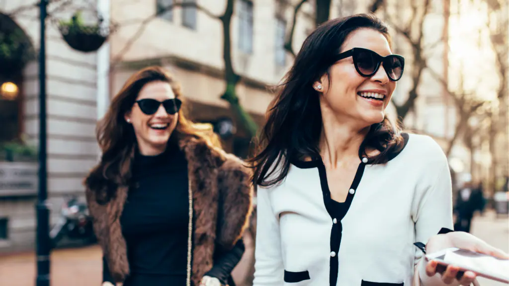 Two women walking on a city street, both wearing sunglasses and smiling.